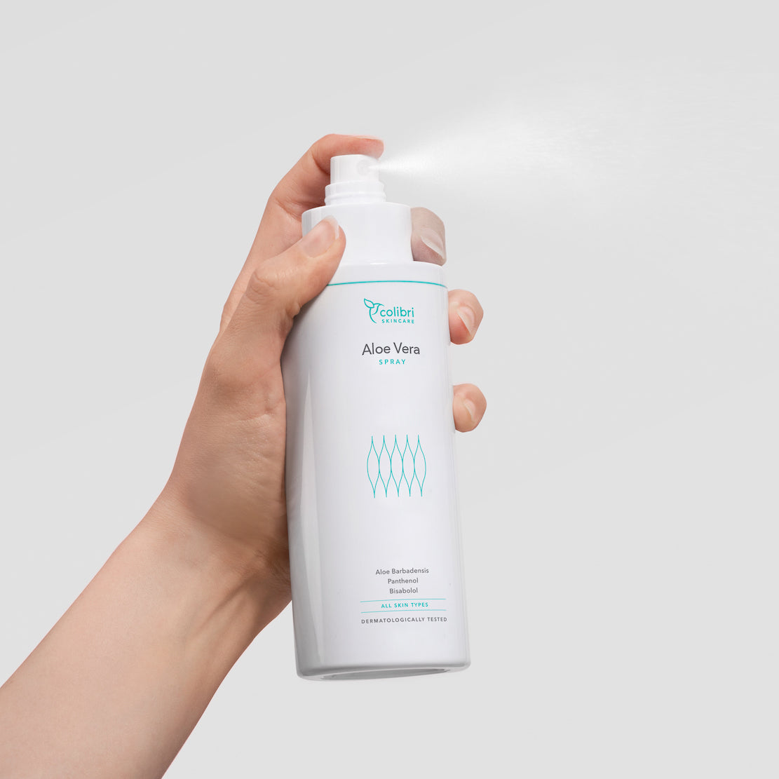 Hand hält eine weiße Flasche Aloe Vera Spray, aus der ein feiner Nebel austritt, vor grauem Hintergrund.