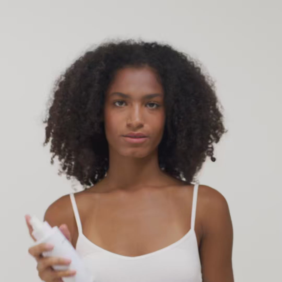 Eine Person mit lockigem Haar hält eine Sprühflasche in der Hand und trägt ein weißes Top vor einem neutralen Hintergrund.