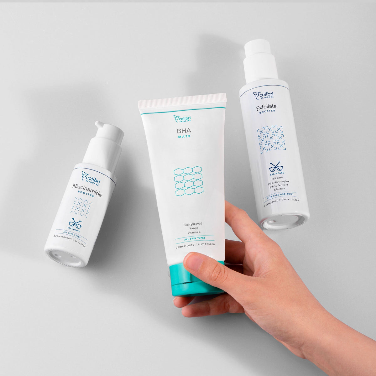 Drei Hautpflegeprodukte auf grauem Hintergrund, eine Hand hält eine BHA-Maske, daneben Niacinamide und Exfoliate Booster.
