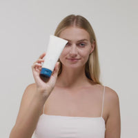 Eine Frau mit blonden Haaren hält eine weiße Tube mit blauer Kappe vor einem neutralen Hintergrund.